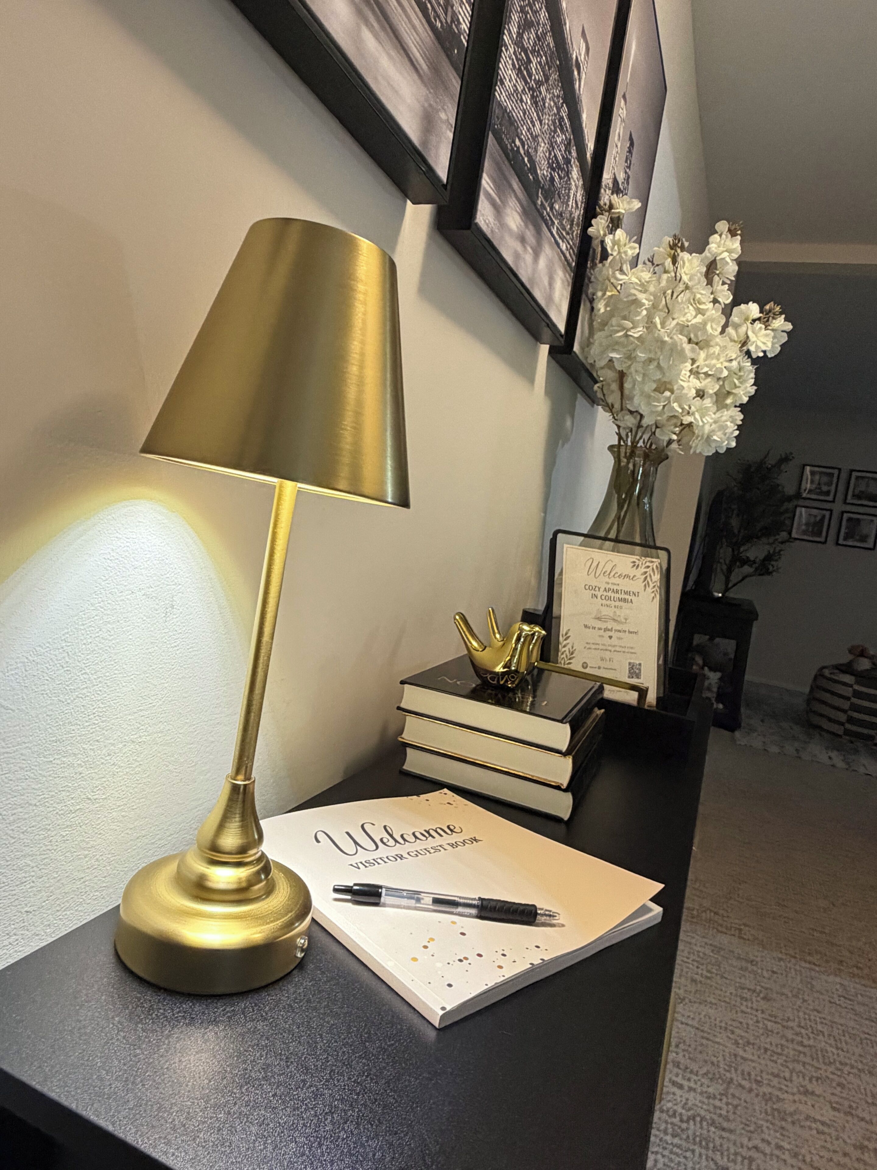 A desk with a lamp, a notebook, a pen, a vase with flowers, and framed pictures on the wall.