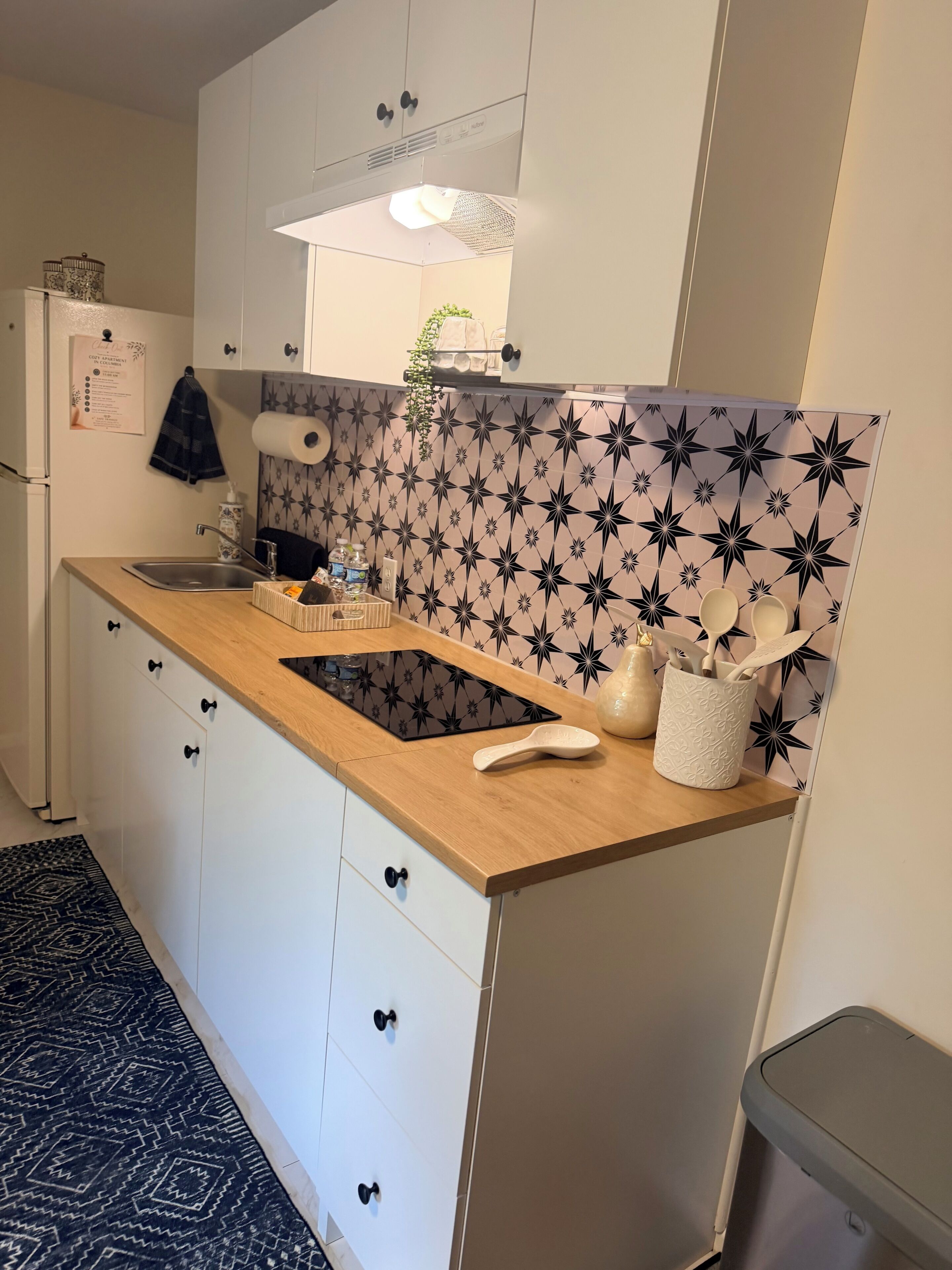 Una cocina con encimera de madera, armarios blancos, salpicadero con estampado de estrellas en blanco y negro, un lavamanos y un cubo de basura.