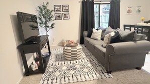 Una sala de estar con un sofá gris, un otoman pequeño y redondo, una mesita auxiliar negra, una planta en maceta y una alfombra estampada.