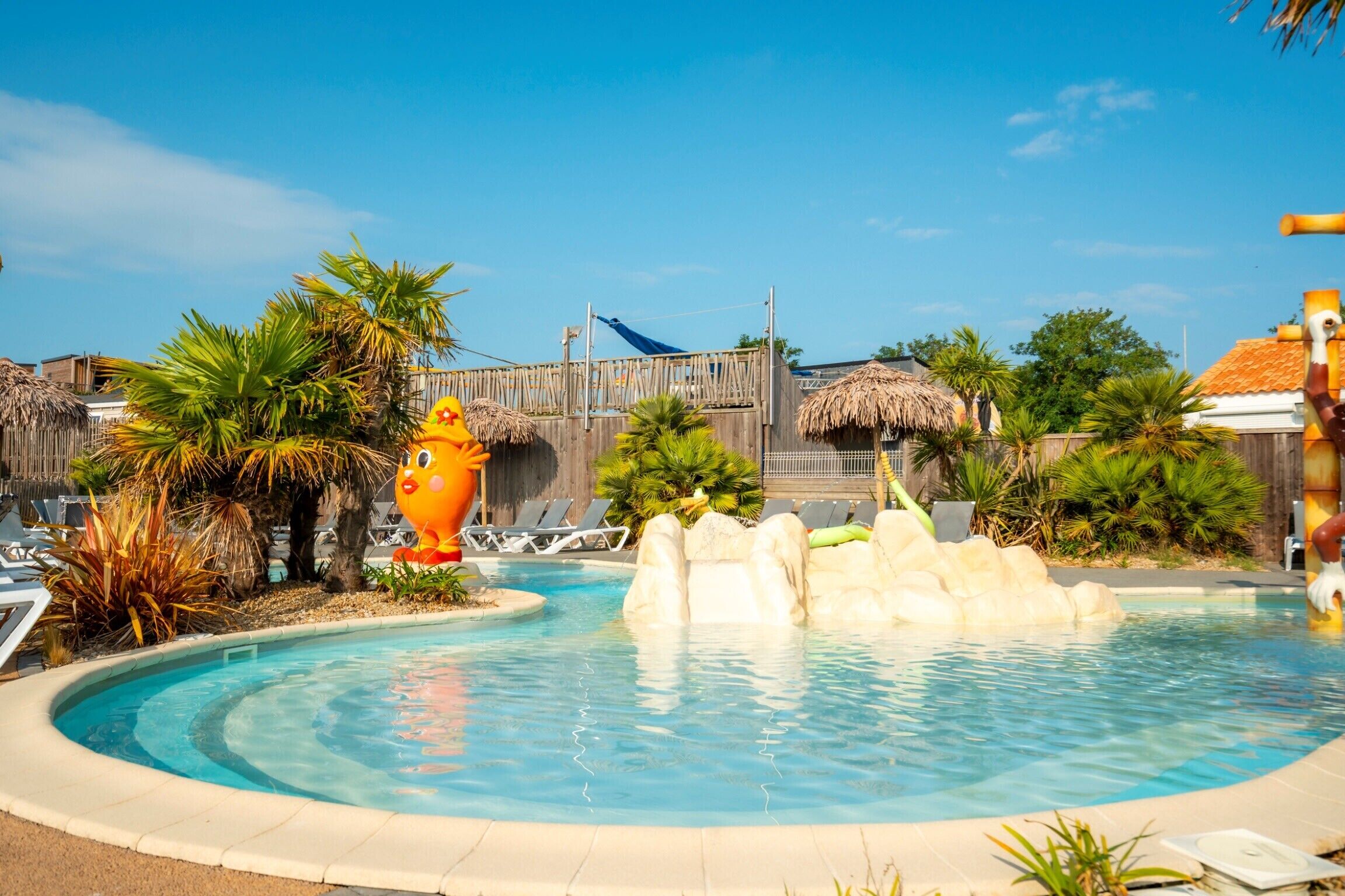 Ein Swimmingpool mit Felsformation, Liegestühlen, Palmen und strohgedeckten Hütten.