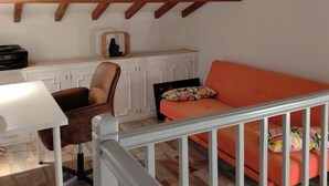 Um quarto estilo loft com vigas de madeira aparentes, teto inclinado, escrivaninha branca, cadeira marrom, sofá laranja e uma pequena estátua em uma prateleira.