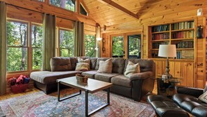Ein gemütliches Blockhaus-Interieur mit Holzbalkendecke, großen Fenstern, einer Couch, einem Couchtisch und einem Bücherregal.