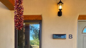 A covered outdoor seating area with wicker chairs, a small table, a hanging chili pepper garland, a wall-mounted mailbox, and a door with a mailbox. - Horse Ranch Casita #B (Santa Fe)