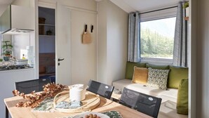 A cozy dining area with a wooden table, chairs, and a plate of lychees. A window with curtains provides a view of trees. - Carva - Confort 3 TV - 6p (3) (La Guérinière)