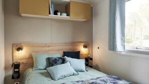 A bedroom with a wooden headboard, two bedside lamps, a bed with a light blue bedspread and multiple pillows, and a window with curtains. - Carva - Confort 3 TV - 6p (3) (La Guérinière)