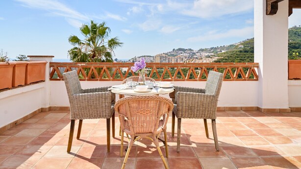 A terrace with wicker chairs and a table, overlooking a cityscape with hills.