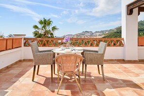 A terrace with wicker chairs and a table, overlooking a cityscape with hills.