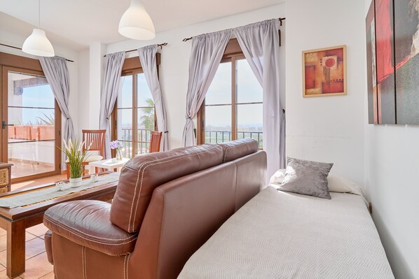 Une chambre à coucher avec un lit, un fauteuil en cuir marron, une table à manger agrémentée d’une plante, et de grandes fenêtres avec des rideaux.