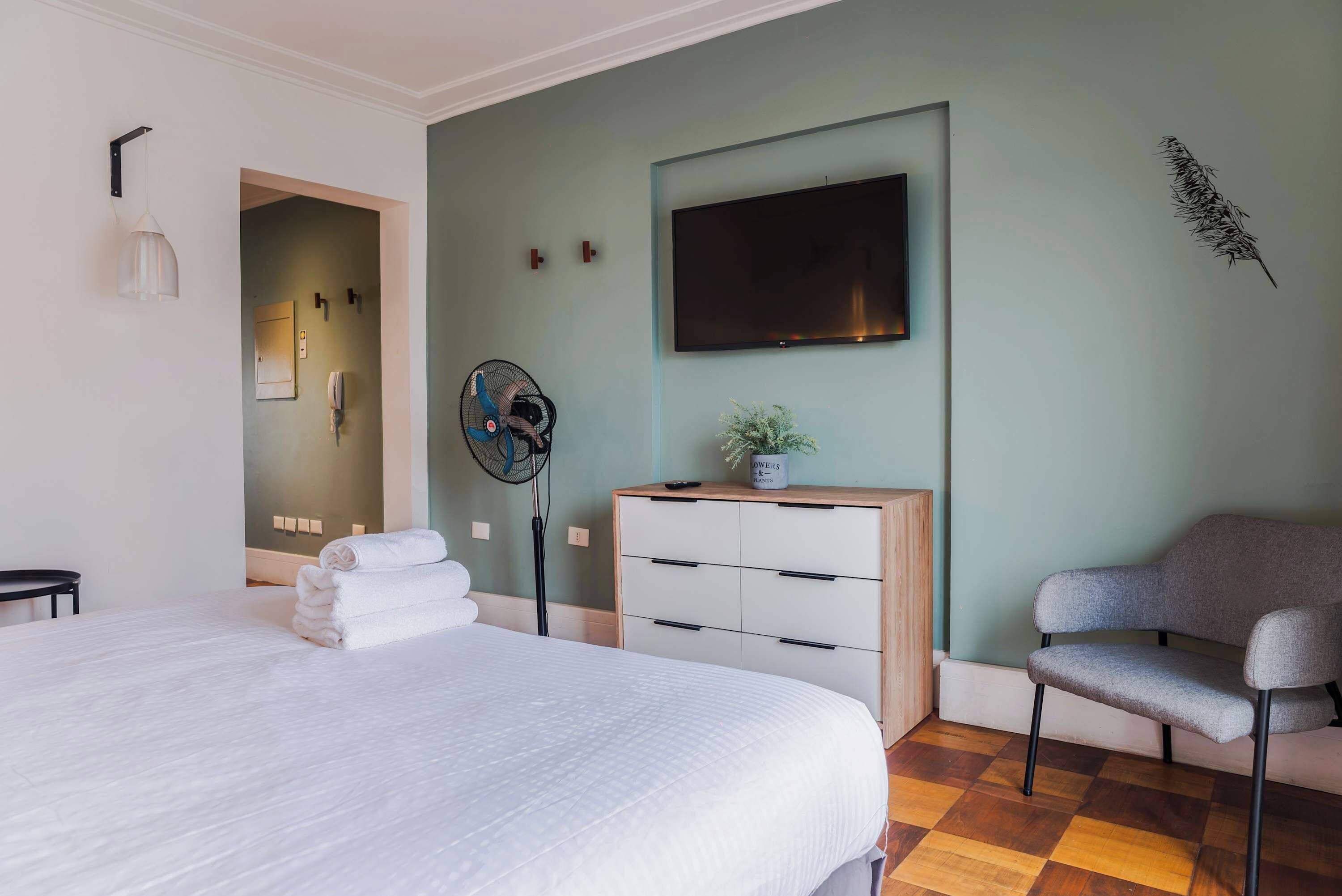 A hotel room with a bed, a desk, a chair, a TV, a fan, and a decorative plant.