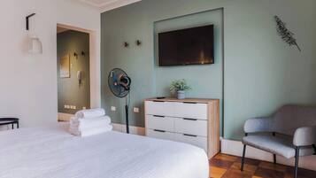 A hotel room with a bed, a desk, a chair, a TV, a fan, and a decorative plant.