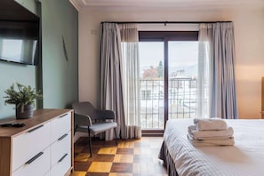 A hotel room with a bed, a desk with drawers, a chair, a potted plant, and a balcony with curtains. - Comfortable Apartment with Full Kitchen and Wifi (Providencia)