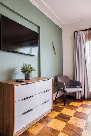 Una habitación con una televisión de pantalla plana montada en la pared, un mueble de madera con cajones, una silla gris y un suelo a cuadros.