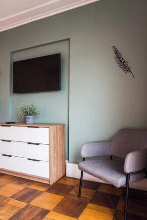 A room with a flat-screen TV mounted on the wall, a wooden cabinet with drawers, a grey upholstered chair, and a checkered wooden floor. - Comfortable Apartment with Full Kitchen and Wifi (Providencia)