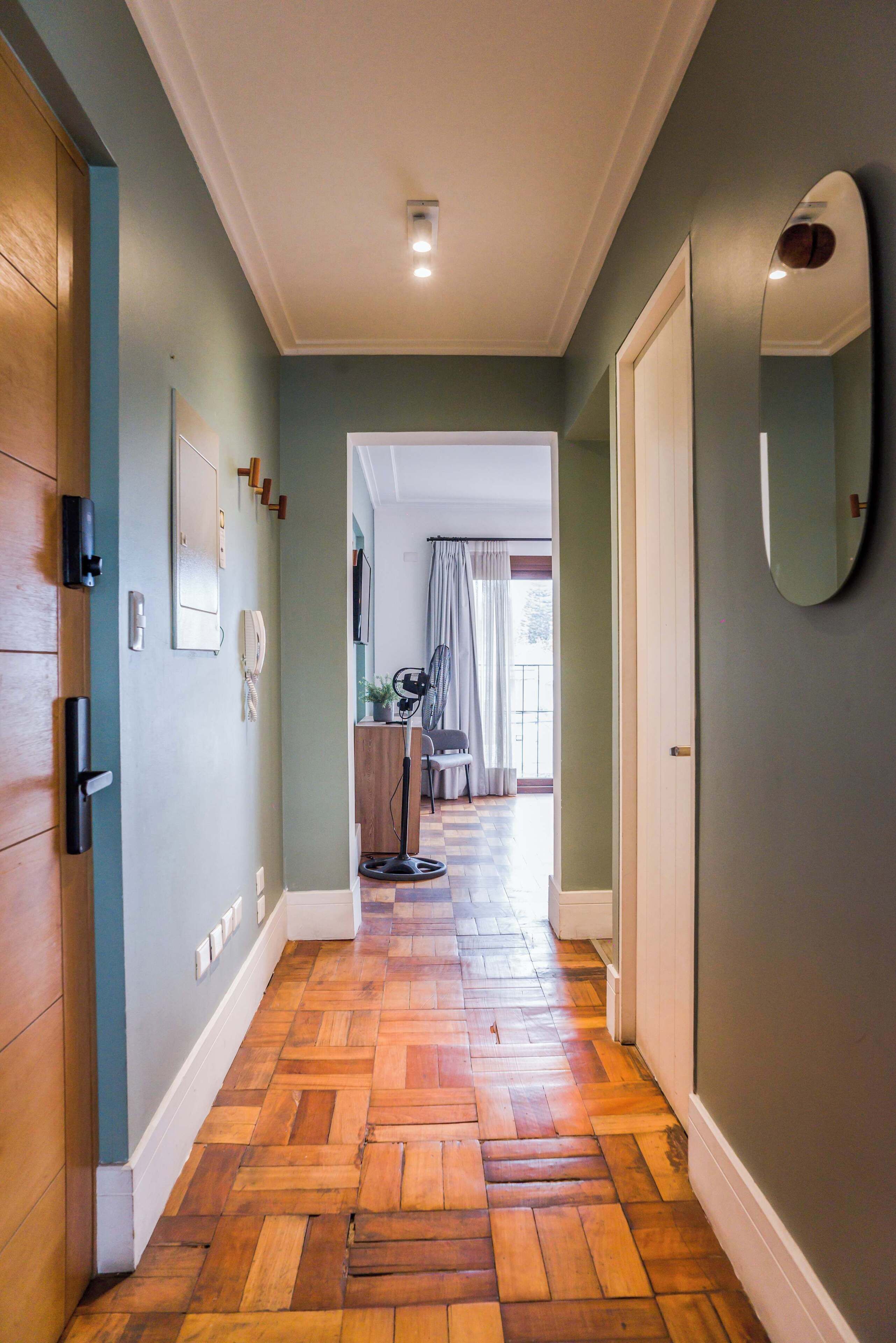 A hallway with wooden flooring, a door on the left, a mirror on the right, and a view into a room with a chair and curtains.