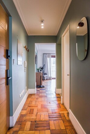 A hallway with wooden flooring, a door on the left, a mirror on the right, and a view into a room with a chair and curtains.