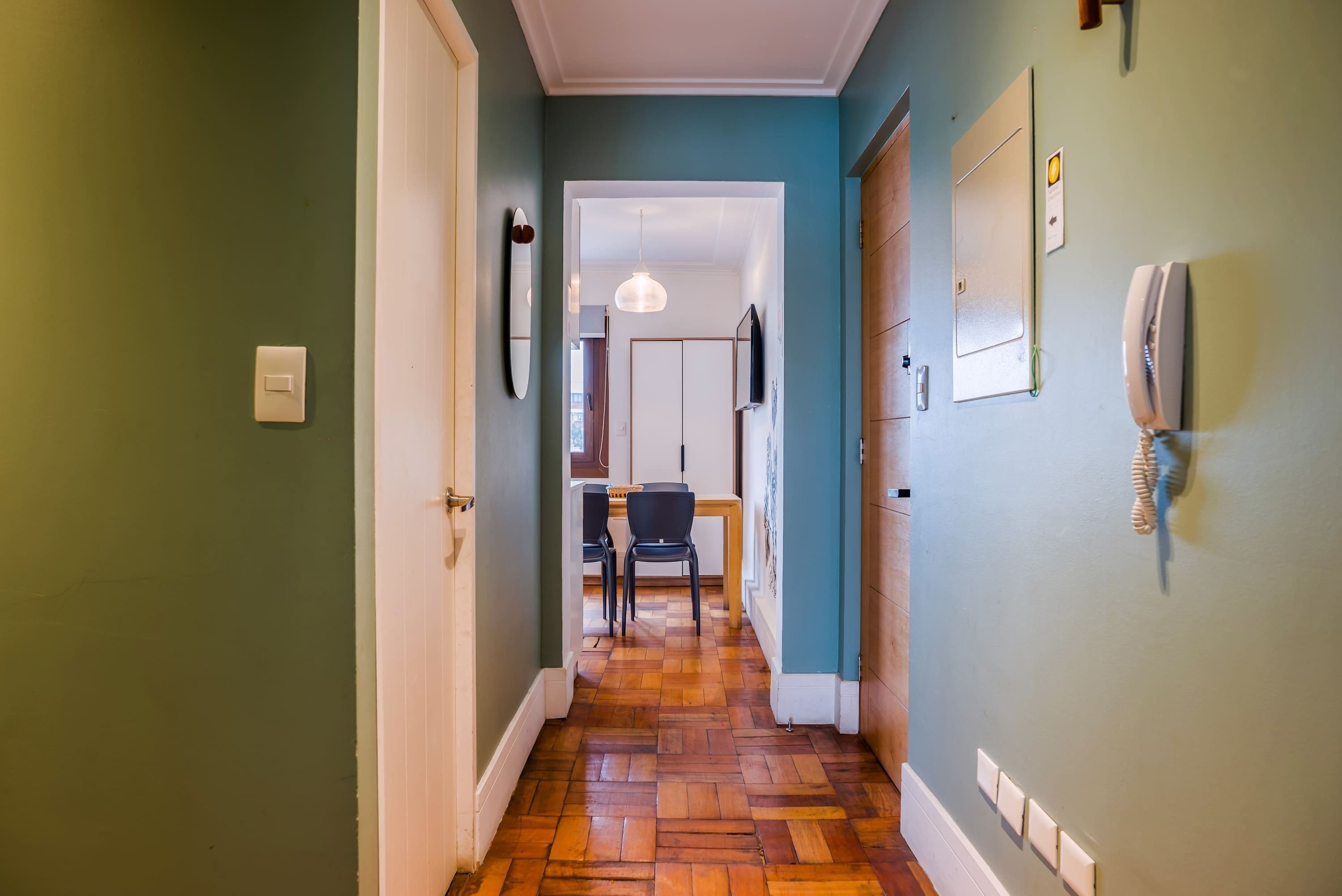 A hallway with a wooden floor, a dining area with chairs and a table, and a wall-mounted telephone.