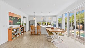 Une cuisine moderne dotée d’un îlot central, d’un coin repas avec des chaises en osier et de grandes fenêtres offrant une vue sur l’extérieur.