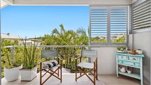 Un balcon avec vue sur des palmiers et un immeuble, comprenant une chaise en bois, une petite table et une commode turquoise agrémentée d’objets décoratifs.