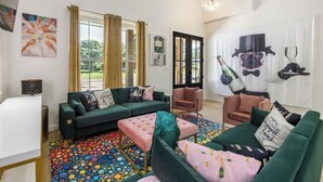 Amplia sala de estar con un sofá verde, un otoman rosa y un sillón rojo. El piso tiene un mosaico colorido y una ventana grande con cortinas.