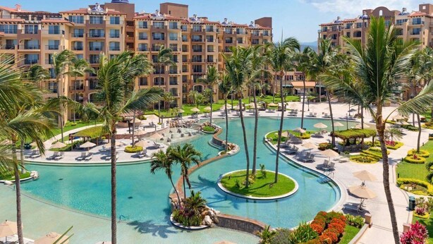 Un resort con una amplia piscina central, palmeras y varios edificios hoteleros al fondo.