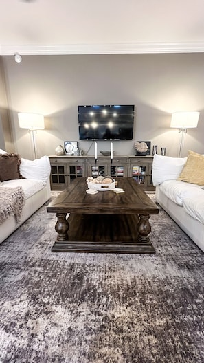 A living room with a dark wooden coffee table, two white sofas with brown and beige cushions, a flat-screen TV mounted on the wall, and two floor lamps.