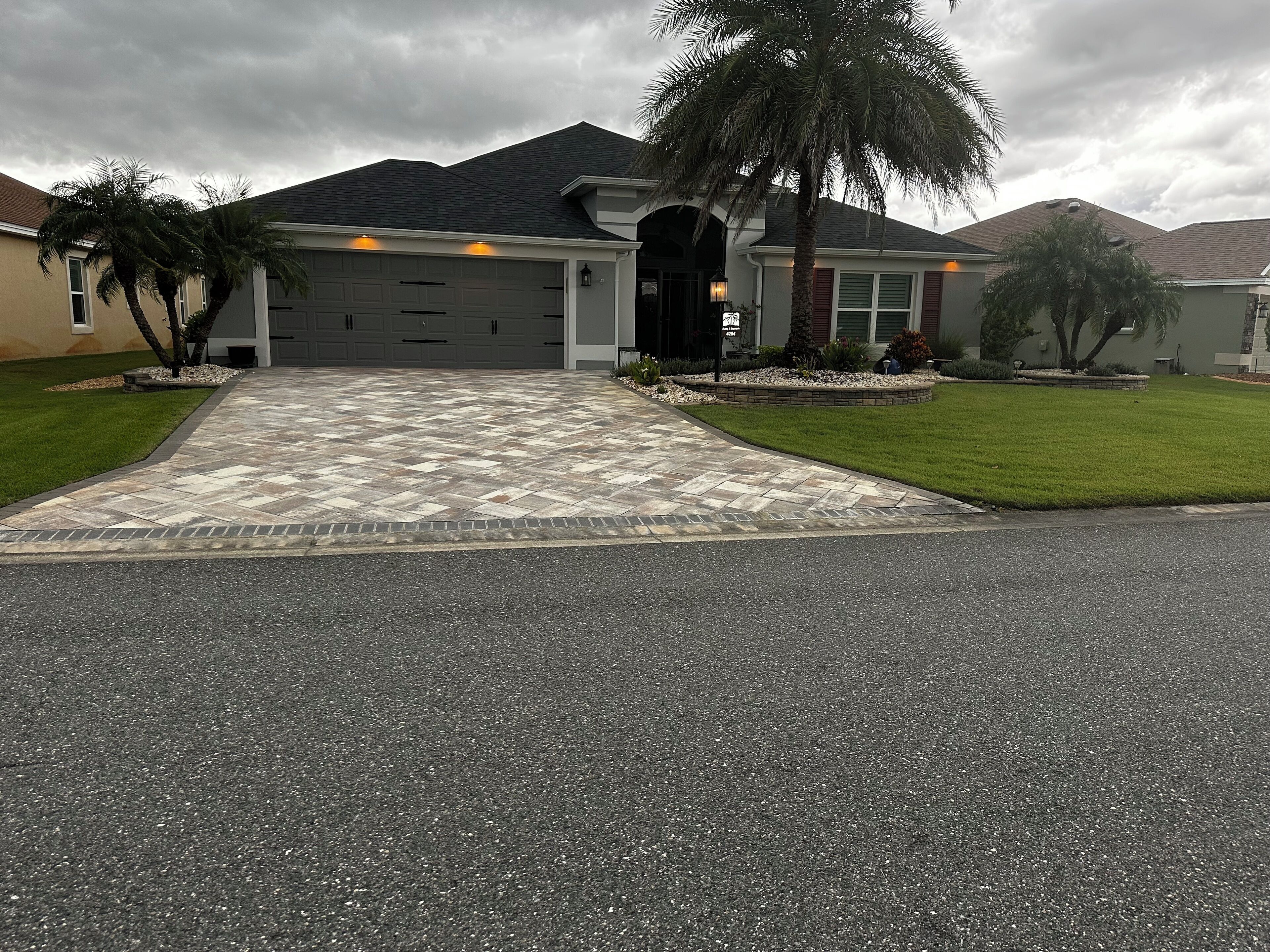 Una casa residencial con un camino de entrada que lleva a un garaje, rodeada de céspedes cuidados y palmeras.