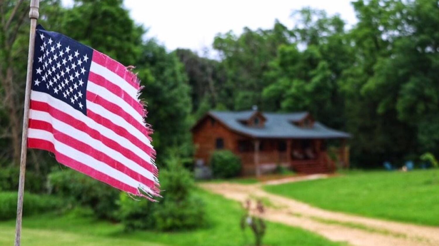 Eine amerikanische Flagge an einem Fahnenmast, im Hintergrund eine Holzhütte.