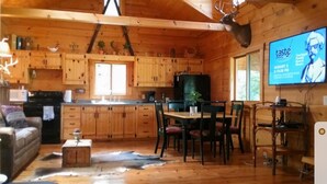 Ein rustikales Blockhaus-Interieur mit Holzwänden, einem Essbereich mit Stühlen und Tisch, einer Küche mit Holzschränken und einem aufgehängten Hirschkopf.