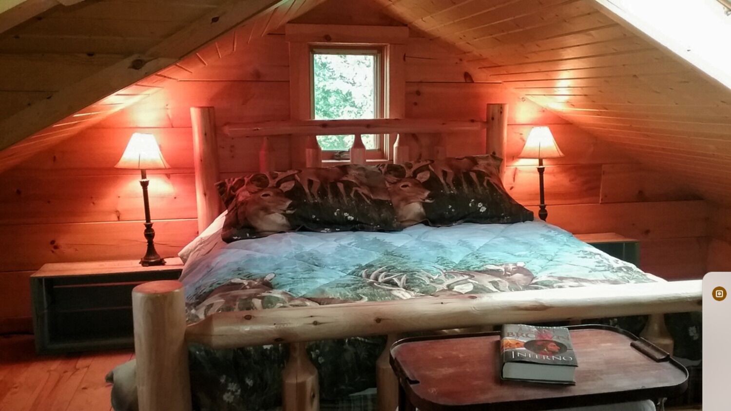A cozy attic bedroom with a wooden sloped ceiling, a bed with deer-themed bedding, two bedside lamps, and a small table with a book.
