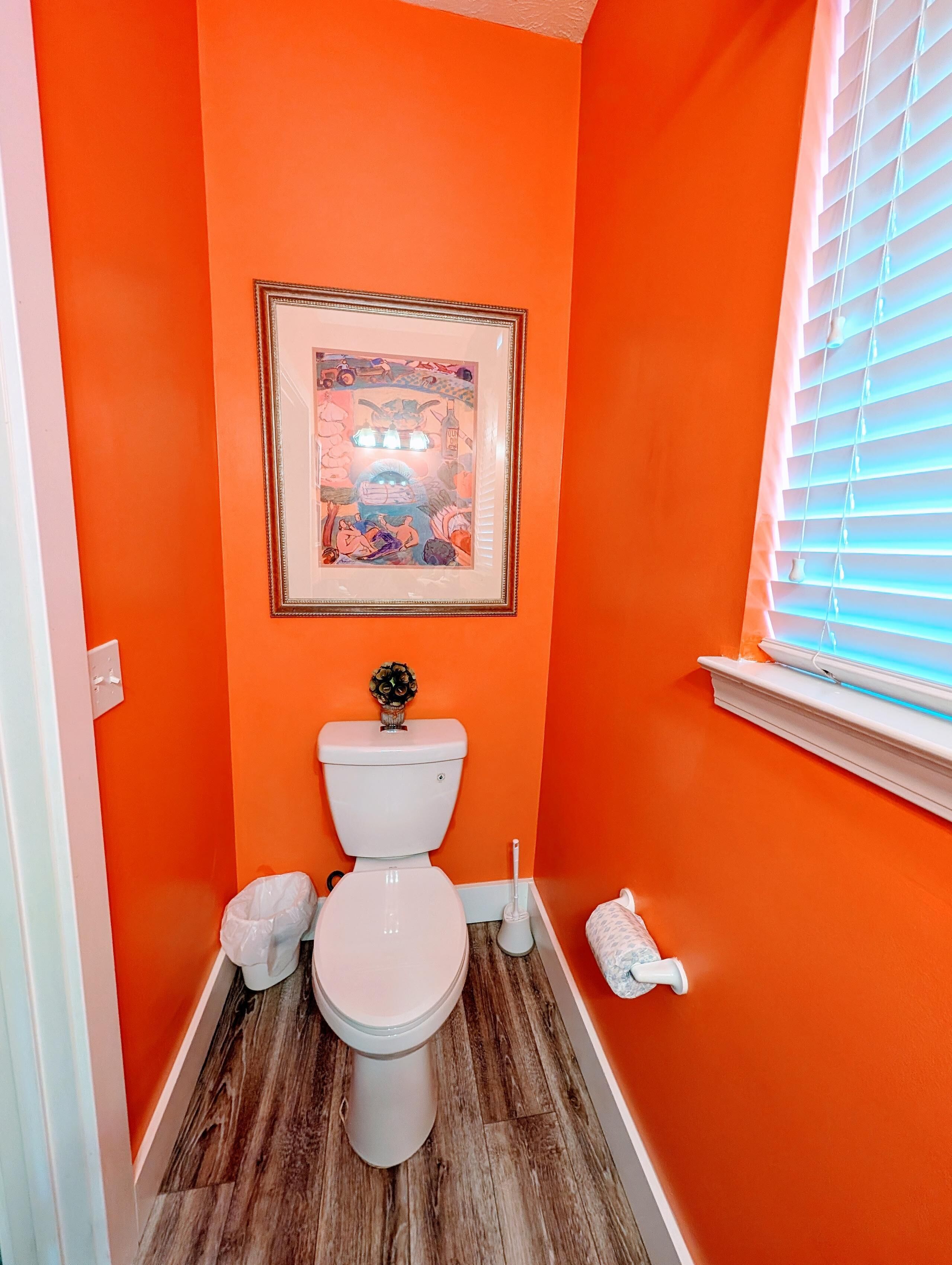 A bathroom with an orange wall, a framed picture, a toilet, a bidet, and a window with blinds.