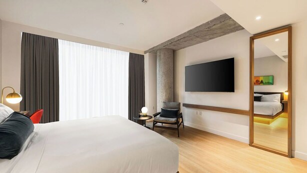 A modern hotel room with a large bed, a flat-screen TV, a chair, a small table with a lamp, and a large mirror. - Arden Hotel (Vancouver)