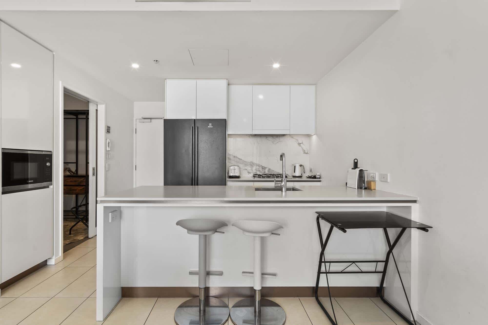 A modern kitchen with white cabinetry, a stainless steel refrigerator, a marble backsplash, and a black bar stool.