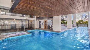 A modern indoor swimming pool with a wooden ceiling, clear blue water, and a view of the cityscape. - Central South Brisbane 2-Bed with Stunning Views (South Brisbane)