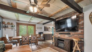 A rustic interior with a wooden ceiling, a fireplace, a TV mounted on a brick wall, and a dining area with wooden furniture. - NEW! NH Getaway l HOT TUB l NearWhiteMountain (Campton)