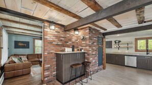 A rustic kitchen with exposed beams, a brick accent wall, and a bar area with stools. - NEW! NH Getaway l HOT TUB l NearWhiteMountain (Campton)