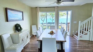 Un espace repas comprenant une table en bois, des chaises à revêtement blanc, un banc blanc et une vue sur l’océan grâce à de grandes fenêtres.