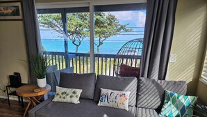 Un canapé gris agrémenté de plusieurs coussins, une petite table en bois sur laquelle est posée une plante en pot, et une vue sur la mer par de grandes fenêtres.