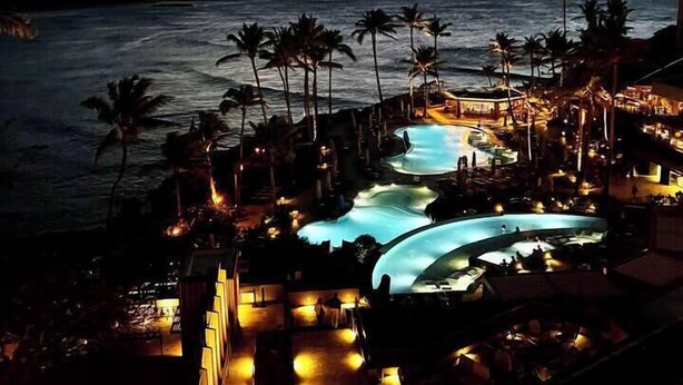 Um resort com várias piscinas, palmeiras e vista para o oceano ao entardecer.