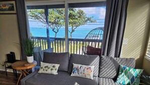 Um sofá cinzento com várias almofadas, uma pequena mesa de madeira com uma planta em vaso e vista para o mar através de grandes janelas.
