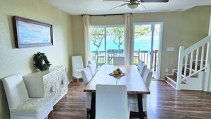Uma sala de jantar com uma mesa de madeira, cadeiras brancas e vista para o oceano através de grandes janelas.