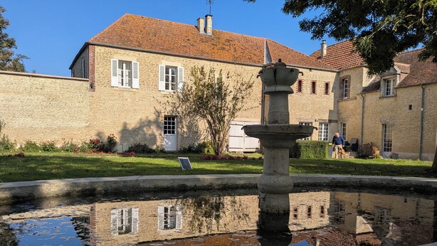 Een gebouw van steen met een pannendak, een fontein op de voorgrond en een heldere weerspiegeling in het water.