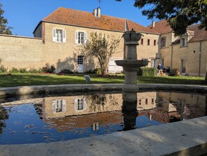Un edificio in pietra con tetto a tegole, una fontana in primo piano e un riflesso limpido nell'acqua.