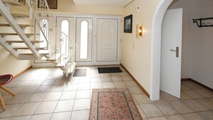 Un pasillo con una escalera blanca, puertas dobles, una silla roja y un tapete estampado.