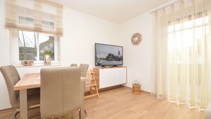 A dining area with a wooden table, chairs, a television displaying a harbor scene, and a window with blinds. - Watthof apartment 11<br>Modern apartment in a quiet location with access to the sea (Stinteck)