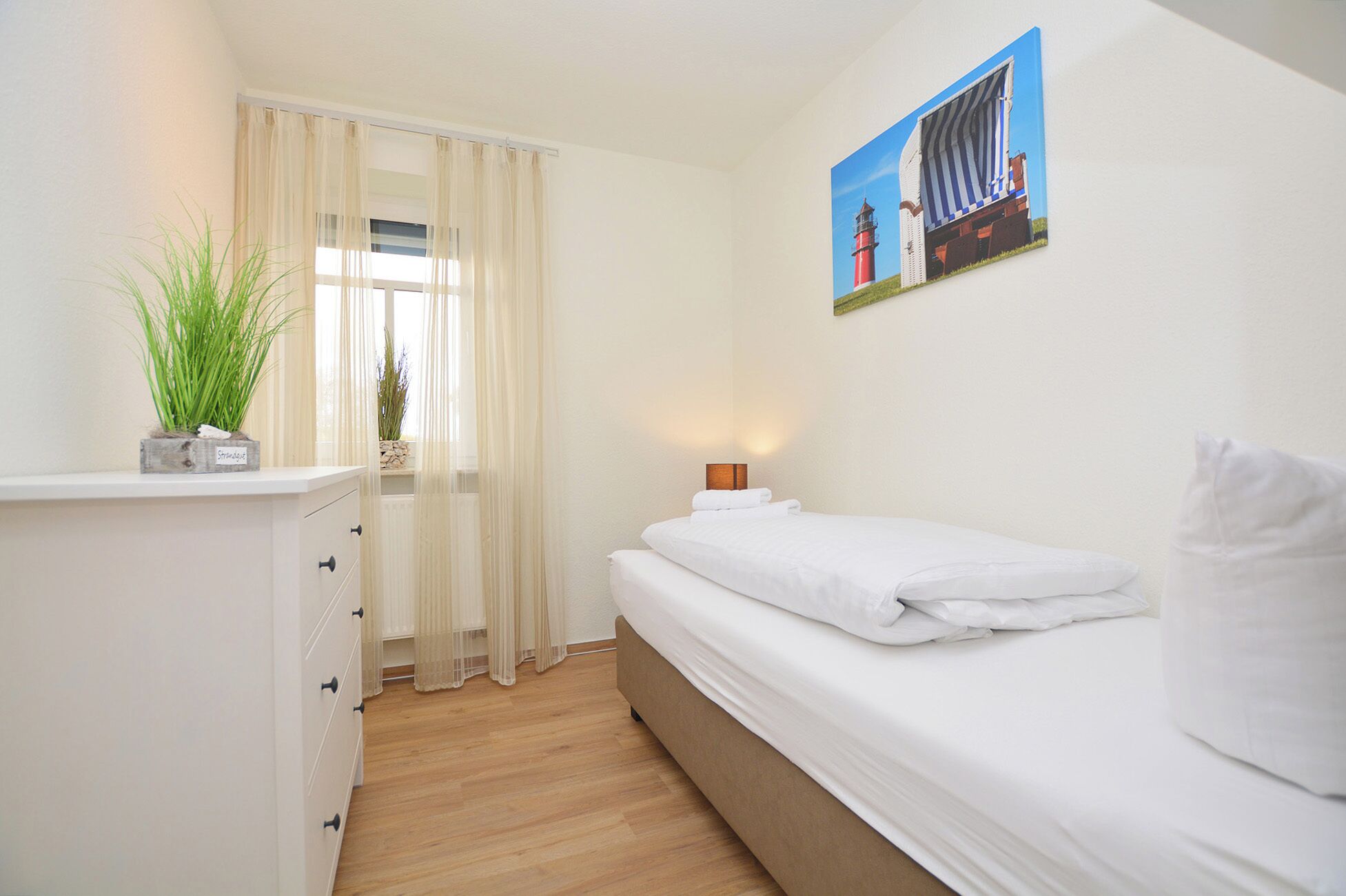 Un dormitorio con cama, mesita de noche, lámpara, ventana con cortinas translúcidas y un cuadro de un faro.