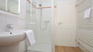 A bathroom with a sink, mirror, shower, and a door leading to another room. - Watthof apartment 13<br>Cozy apartment with terrace and access to the sea time (Stinteck)