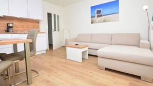 A modern living room with a sofa, a dining table, and a kitchen area with cabinets and a coffee machine. - Watthof apartment 13<br>Cozy apartment with terrace and access to the sea time (Stinteck)