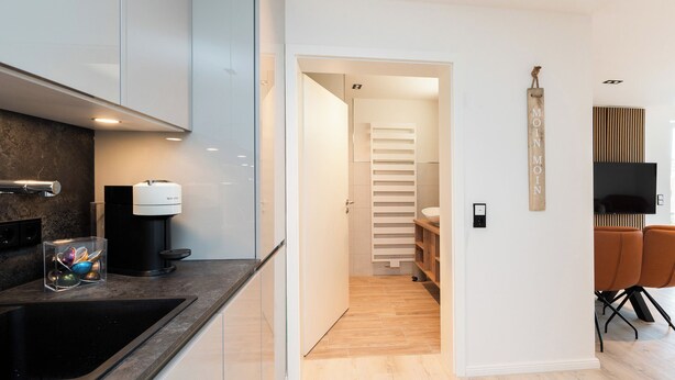 A modern kitchen with a built-in stove, a coffee machine, and a sink. There is a wooden floor and a white wall with a mounted TV. - Inseltörn apartment 10 (Büsum)