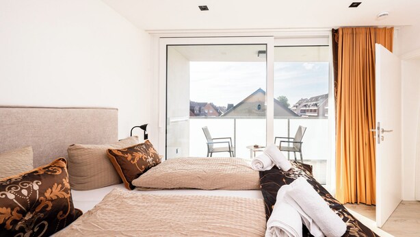 A hotel room with a bed, a sofa, a balcony with a table and chairs, and a door with curtains. - Inseltörn apartment 6 - Comfortable for 6 people & admission to the swimming pool included. (Büsum)