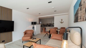 A modern living room with a sofa, armchairs, a coffee table, and a TV. There is a dining area with a table and chairs, and a kitchen in the background. - Inseltörn apartment 6 - Comfortable for 6 people & admission to the swimming pool included. (Büsum)
